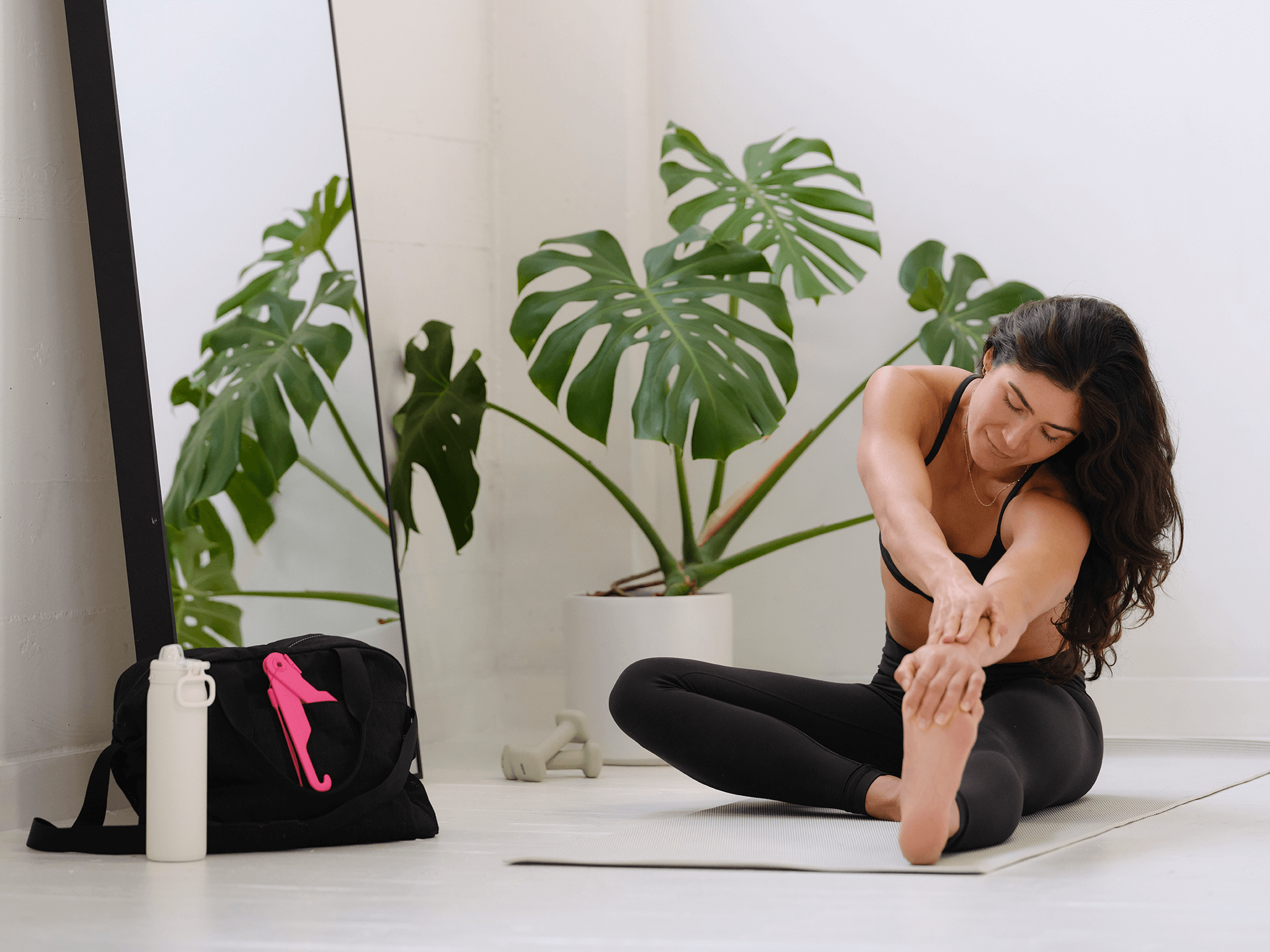 Woman stretching on a yoga mat in a studio with plants and a gym bag with a pink BraClaw tool.